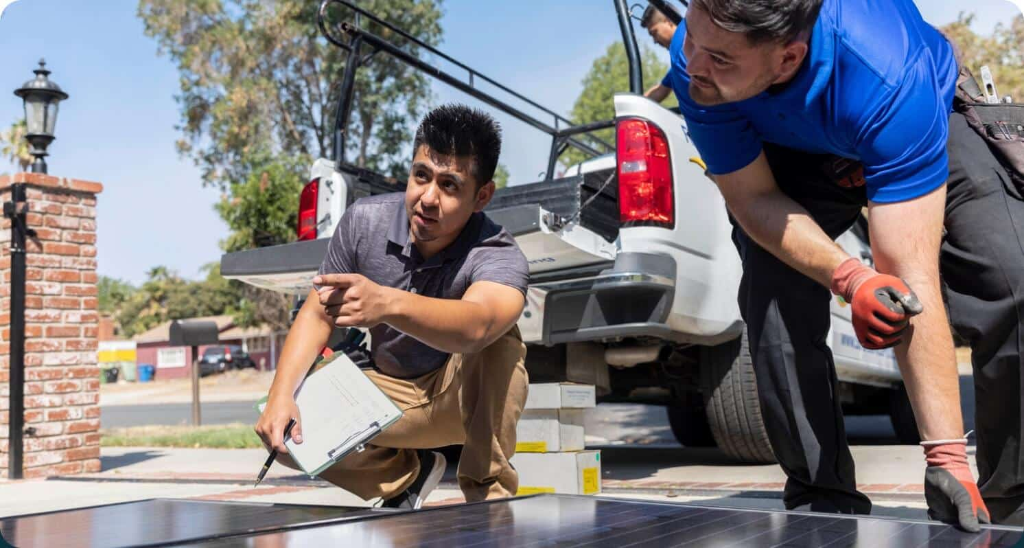 A team of three people working outside taking supplies off of a truck. One person is in the truck and the other two are on the ground. One man is lifting a large panel wile the other is pointing to the left and holding a clipboard.